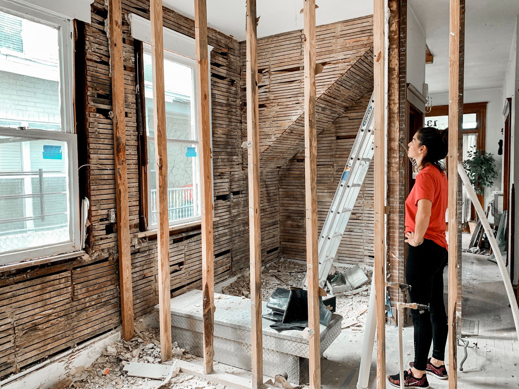 A woman standing in a room stripped to its wood studs and lath, with a ladder against the wall.